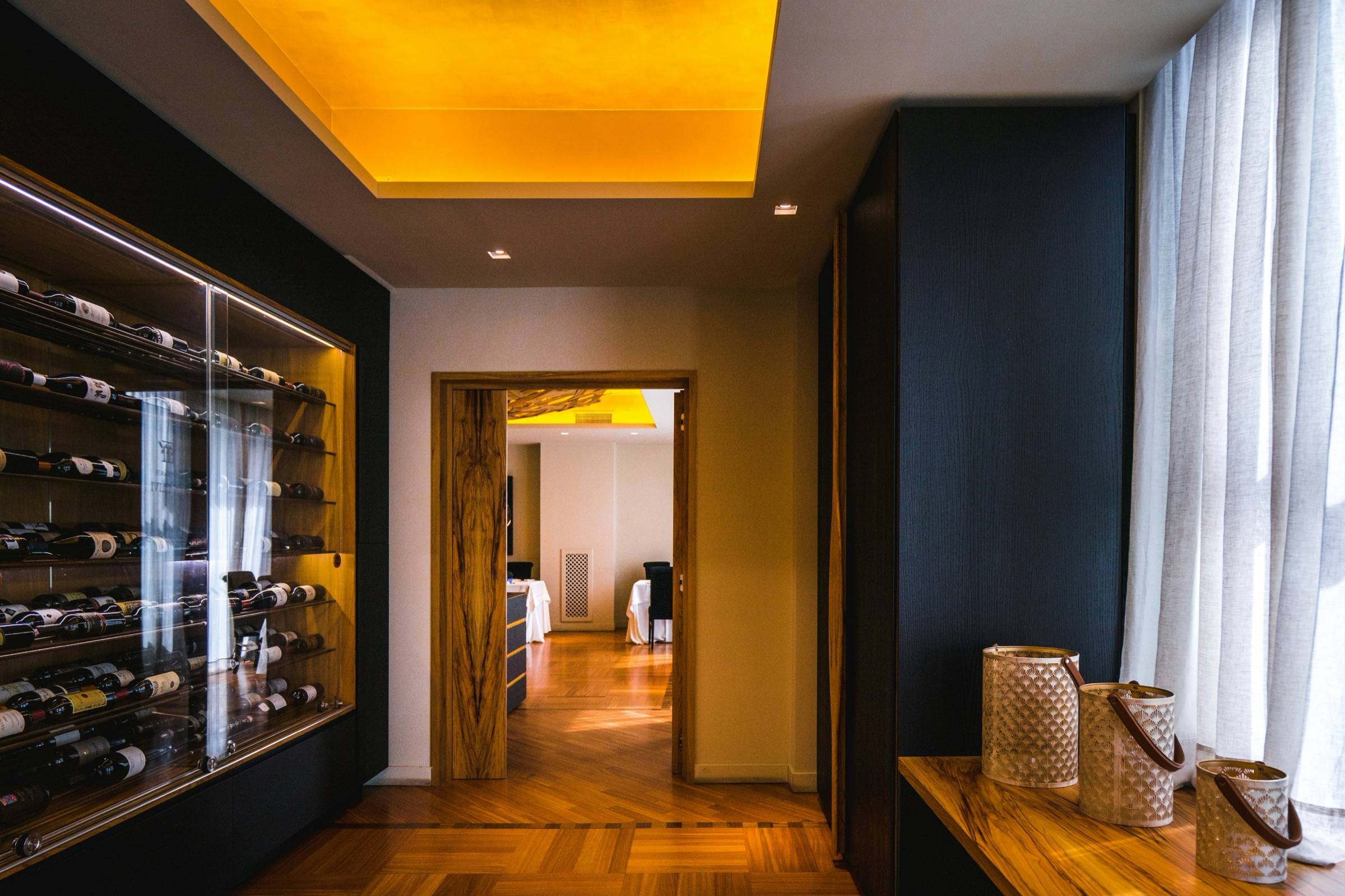 Glass-enclosed wine room with lit racking set into a wall along a modern hallway with wood floors